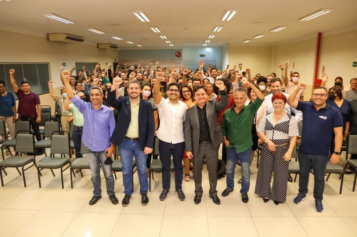 Secretários posicionados à frente do auditório, com as mãos levantadas